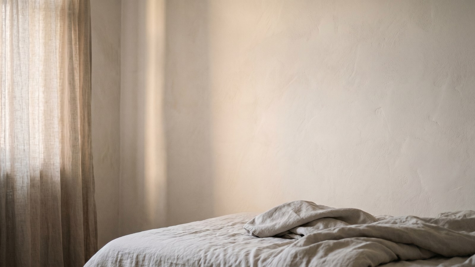 Soft dawn light through a gauzy raw-linen curtain falling across a hand-trowelled lime-plaster wall and the rumpled corner of an unmade bed in undyed linen.
