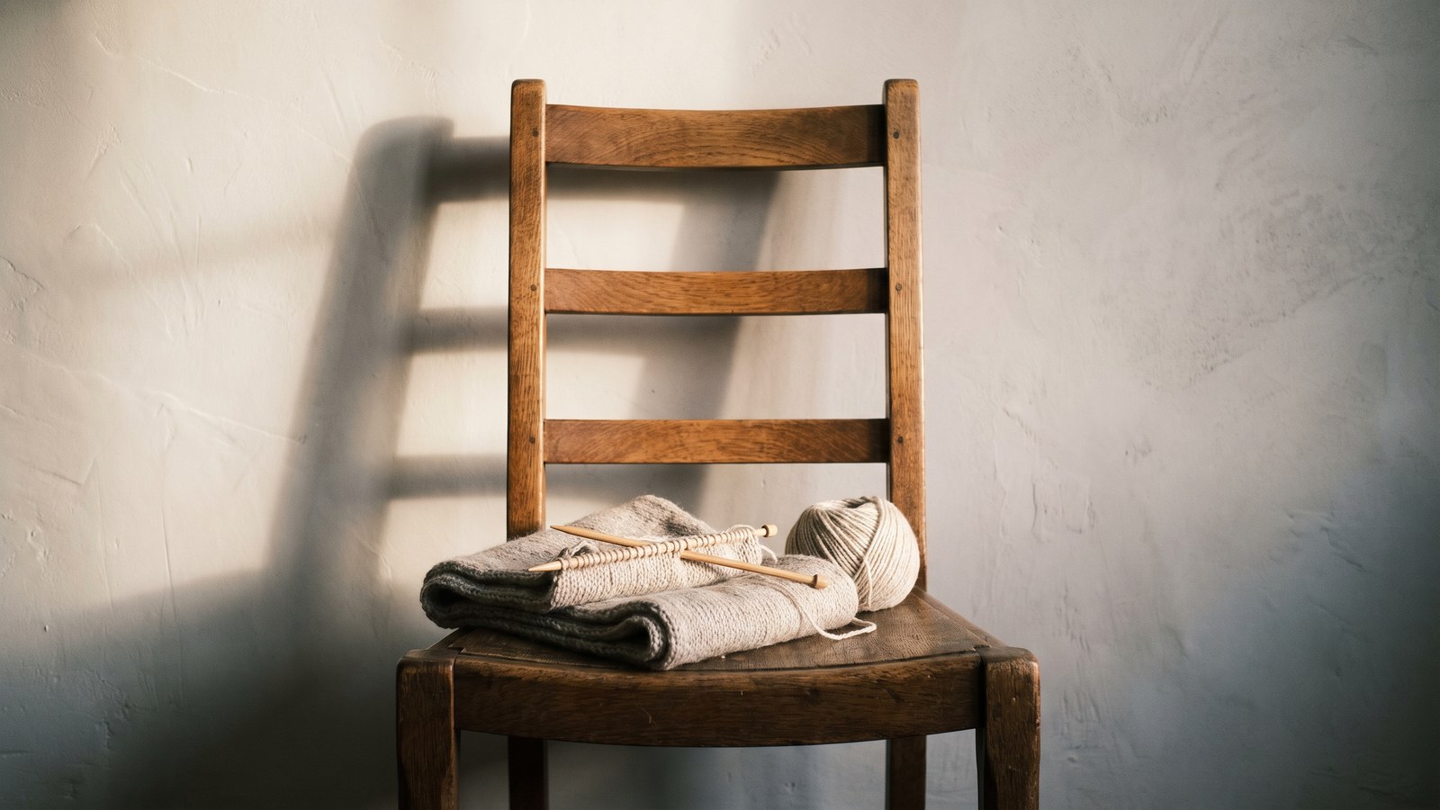 A half-finished scarf in undyed oatmeal wool folded on the seat of an old ladder-back oak chair, two wooden needles still in the row and the ball of wool beside it, morning light throwing the chair's shadow across a lime-plaster wall.