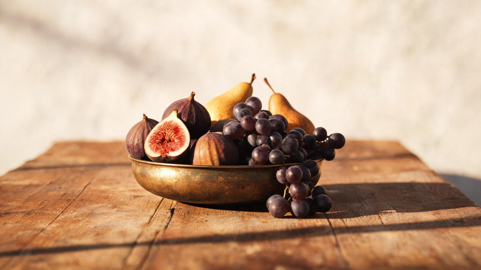 A footed aged-brass compote generously filled with ripe autumn fruit, figs with one split to show its red interior, warm-skinned pears, and a cluster of dark grapes spilling over the rim, set on a worn wooden table in soft golden morning light against a bare lime-plaster wall.