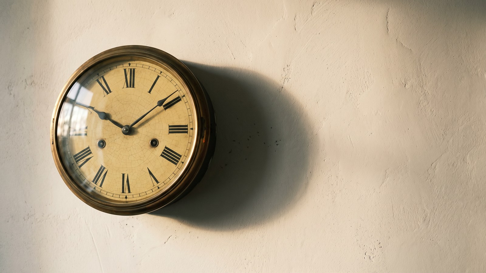 An old brass-bezelled wall clock hanging alone on a bare lime-plaster wall, its pale enamel dial yellowed and finely crazed with age, casting a long soft shadow in low morning light.